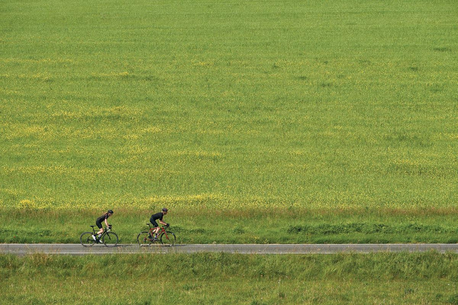 Two cyclists riding a Orro carbon road bike, cycling on a small flat road next to a big green field